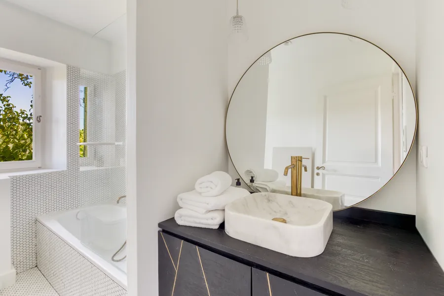A bright bathroom with a large round brass mirror and marble basin